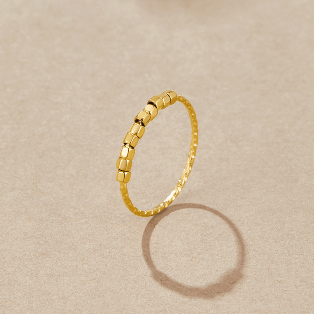 Alison Fidget Ring, standing upright on a beige surface, casting a shadow; gold band with cube beads