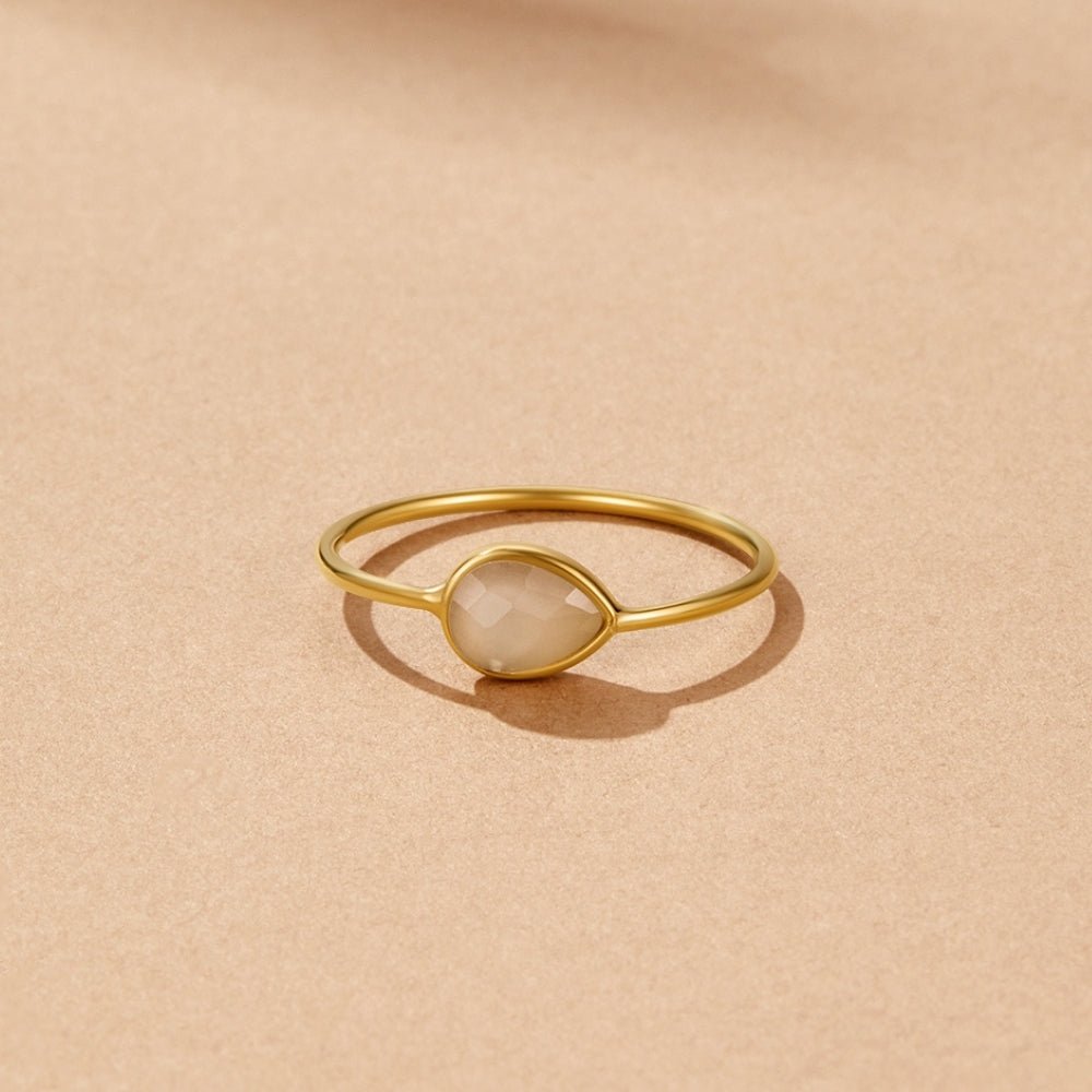 Clara Teardrop Stone Ring, gold ring with a pale teardrop stone, lying on sand-coloured surface