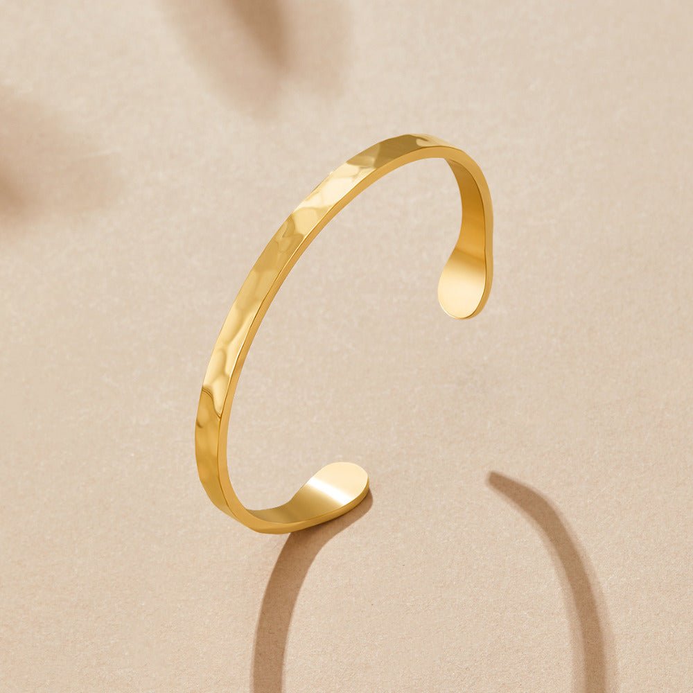 Gold Alexa Bangle, open cuff bangle lying on a beige surface, hammered gold texture
