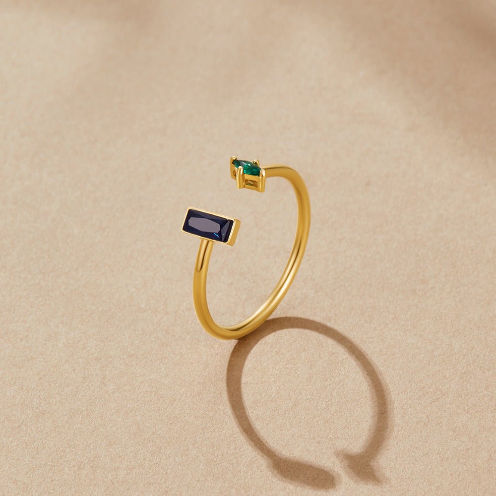 Gold Amore Crystal Ring, open gold ring on beige surface with navy and green baguette stones