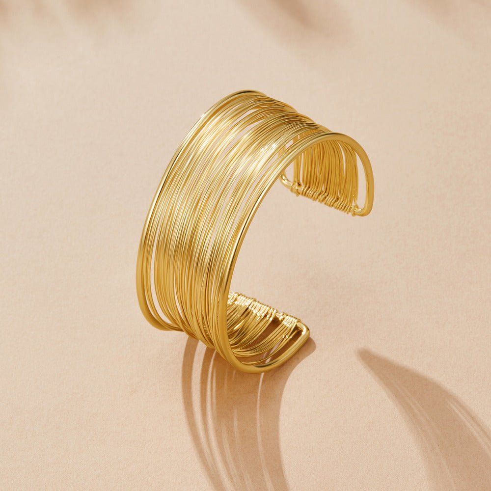 Gold Leona Wide Cuff Bangle, standing open on a beige surface, layered gold wires with shadow
