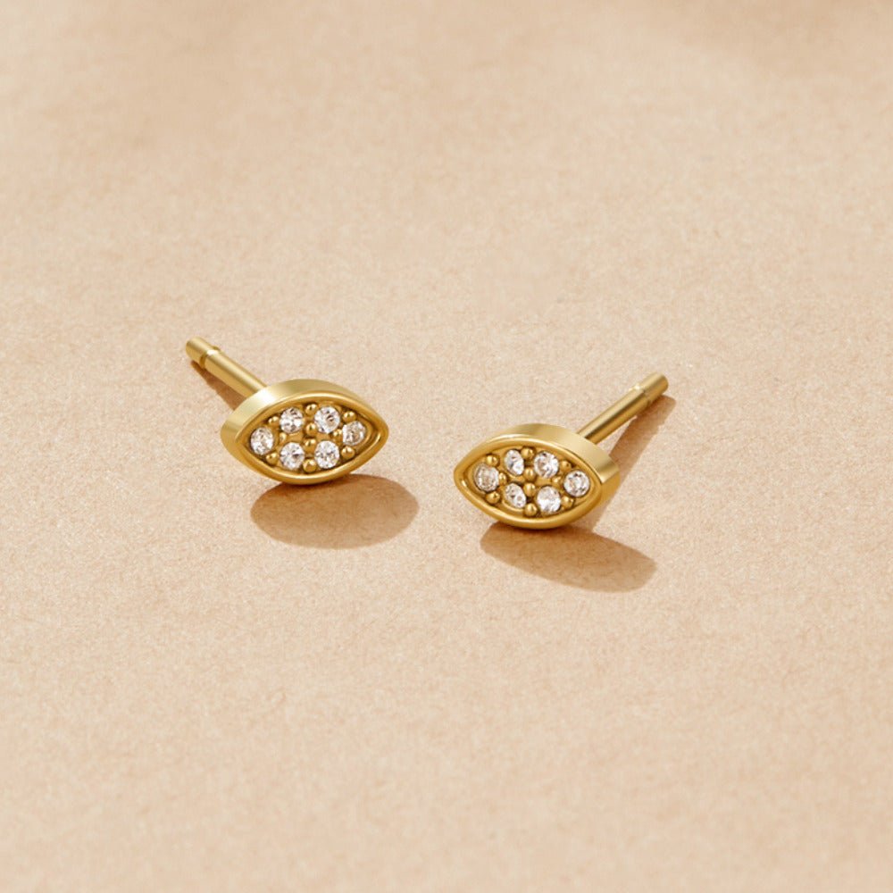 Gold Montana Stud Earrings, a pair of almond-shaped gold studs set with small clear stones, on a beige surface