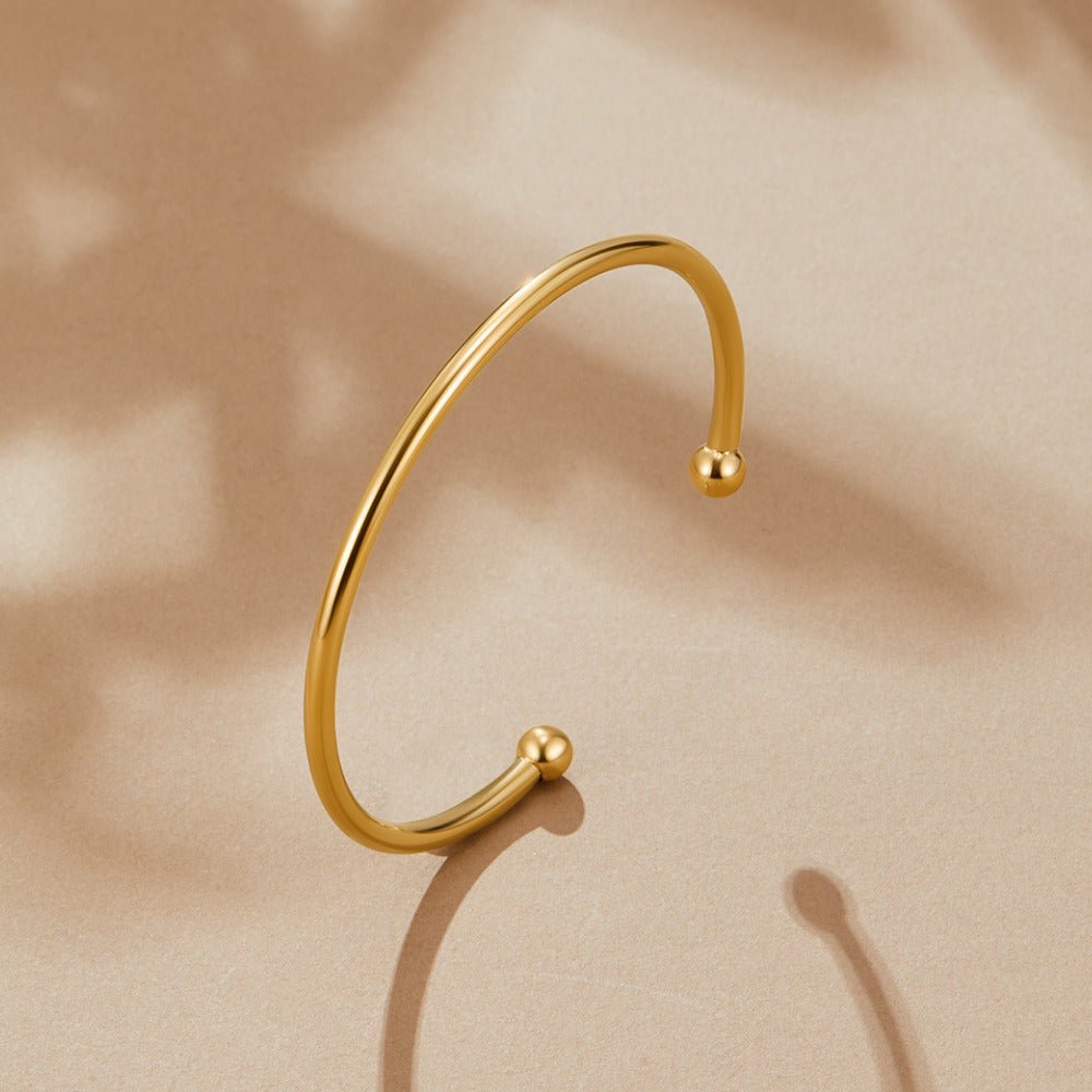 Gold Penelope Bangle, open gold cuff bangle with ball ends, lying on beige surface with leaf shadows