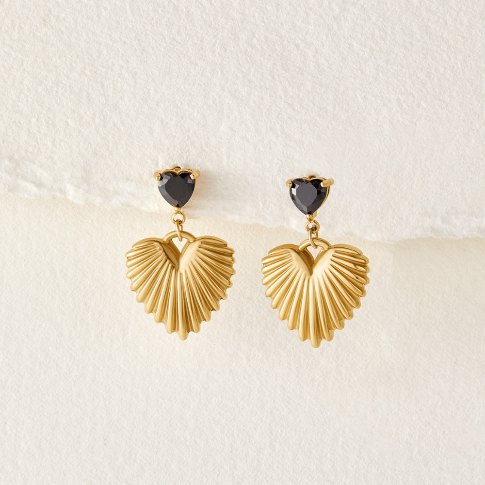 Hera Gold Heart Drop Earrings, a pair of drop earrings laid flat on textured cream paper