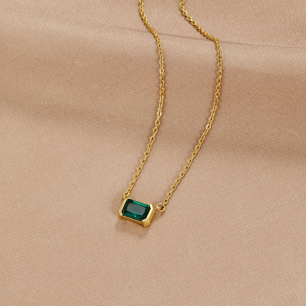 Monica Green Emerald Necklace, laid flat on beige satin, gold chain with green emerald-cut pendant