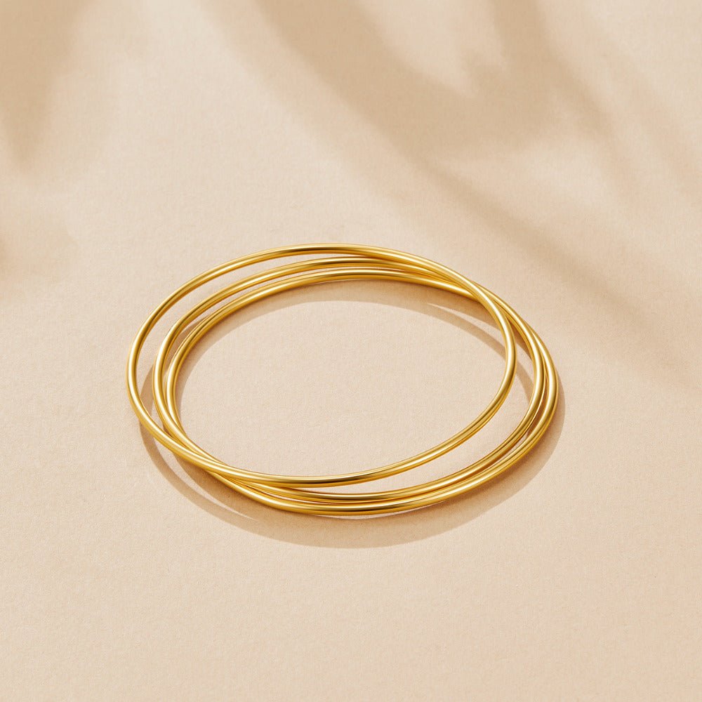 Solana Gold Bangle Set, three thin gold bangles stacked loosely on a beige surface