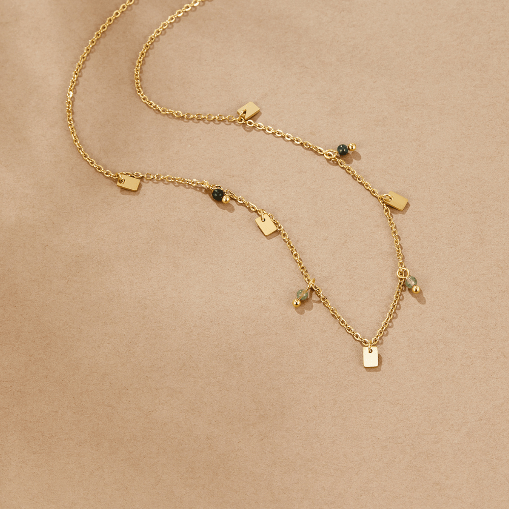 Tara Gold Square Droplet Necklace, laid flat on beige paper, gold chain with dark green beads