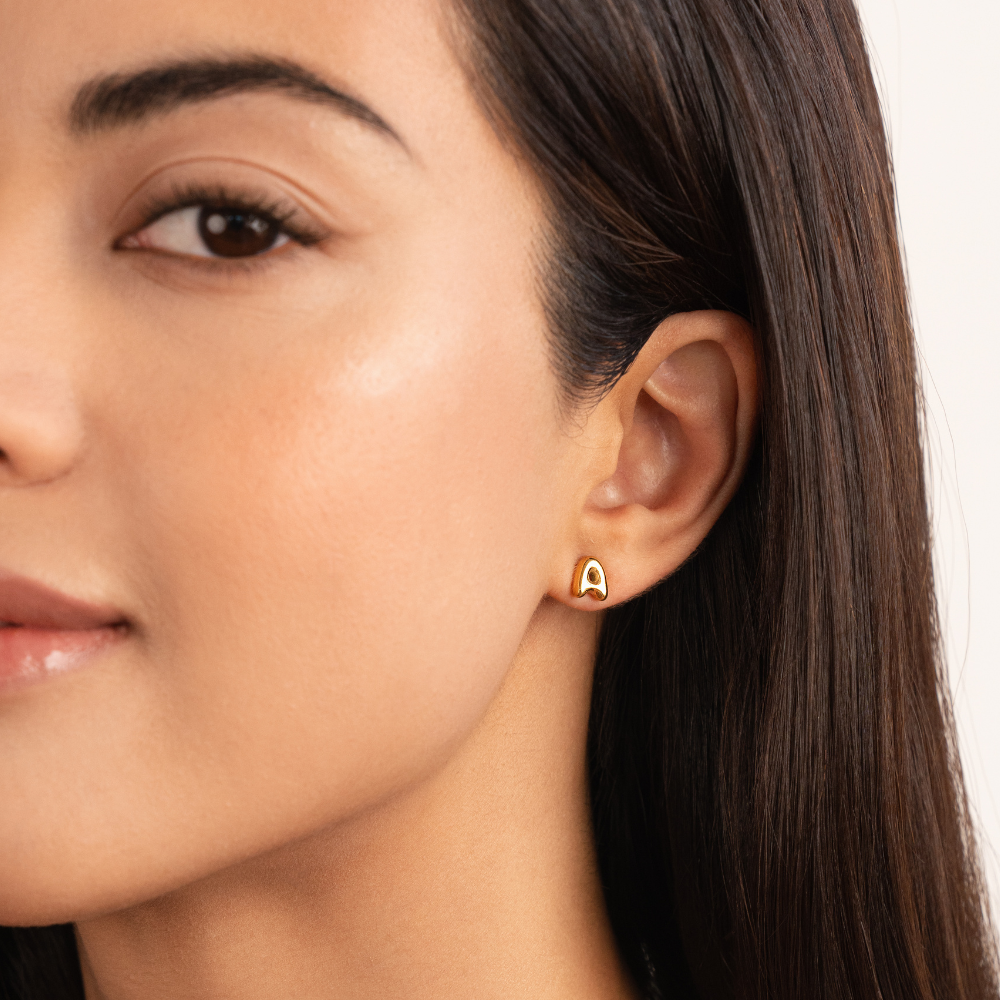 Gold Bubble Letter Earrings, worn on an ear by a model with long dark hair, close-up profile