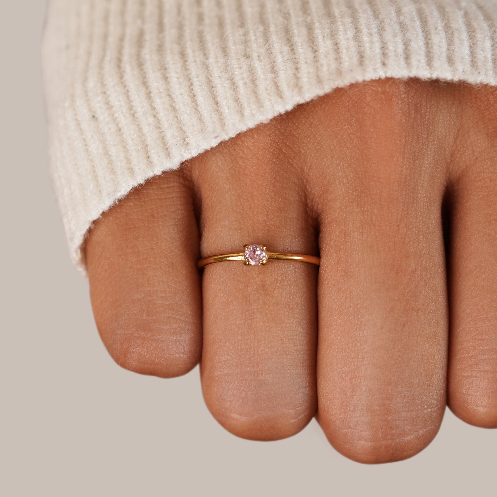 Birthstone 18K Gold Rings, worn on a finger with a cream knit sleeve, pink stone in gold