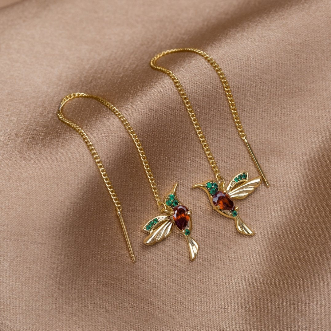 Crystal Hummingbird Tassel Earrings, a pair of gold hummingbird earrings on a tan satin cloth