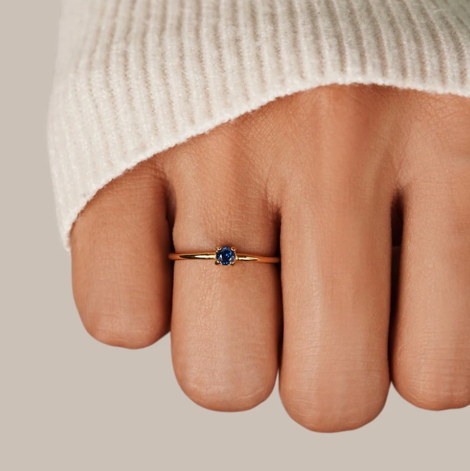 December Birthstone 18K Gold Ring, worn on a finger with a cream knit sleeve, blue stone close-up
