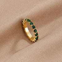 Emerald Beauty 18K Crystal Ring, gold eternity ring with green stones, resting on beige fabric