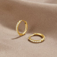 Eternity Hoop Earrings, two gold hoops with a row of clear stones, lying on beige fabric
