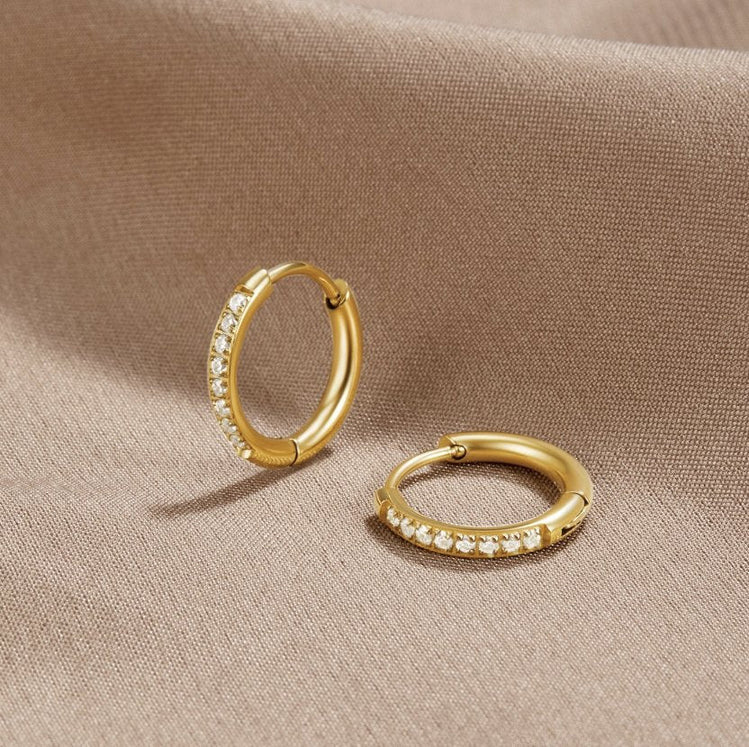 Eternity Hoop Earrings, two gold hoops with a row of clear stones, lying on beige fabric
