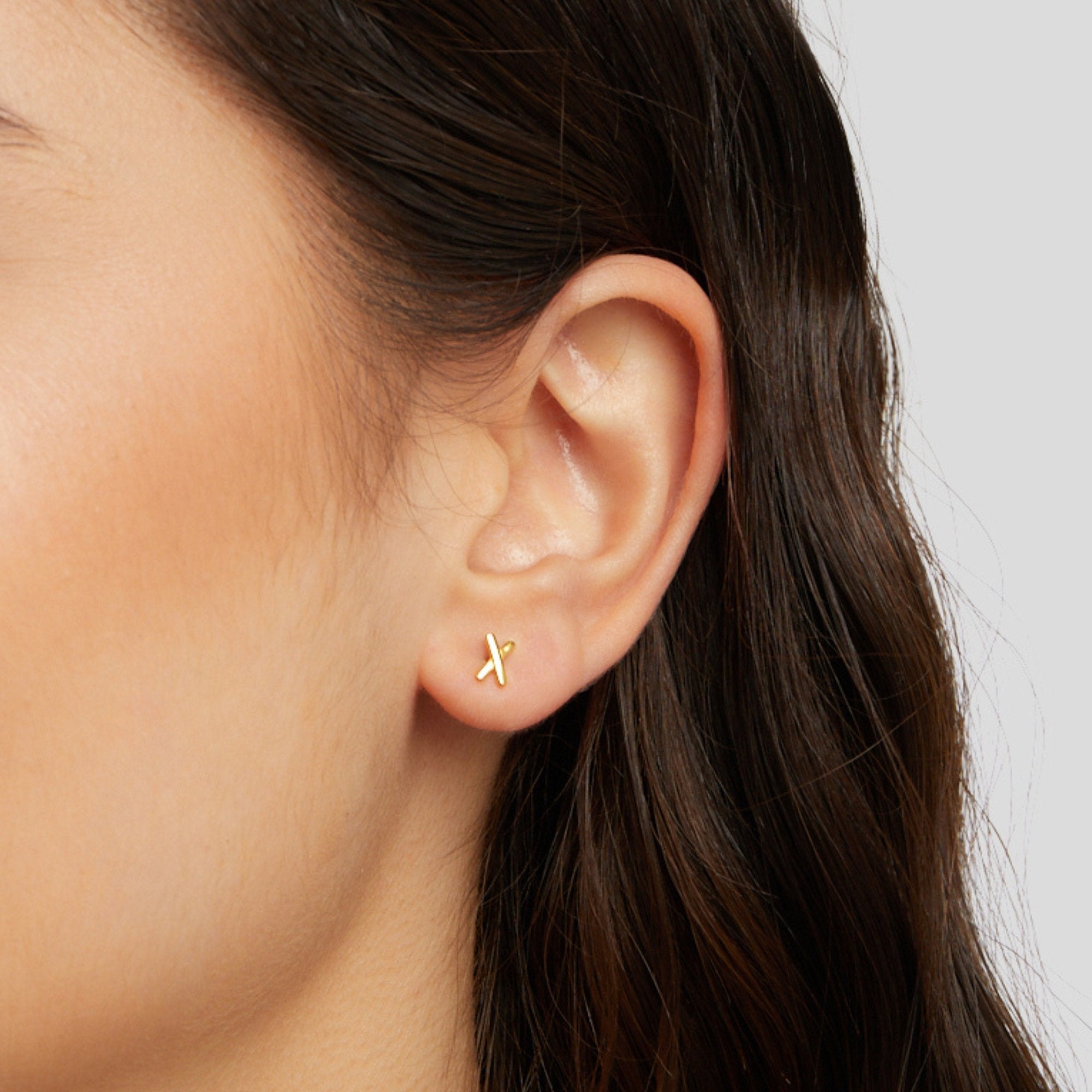 Graffiti X Stud Earrings, worn on an ear, small gold cross-shaped stud