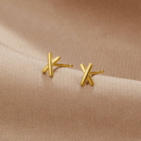 Graffiti X Stud Earrings, pair of gold X-shaped stud earrings on beige fabric