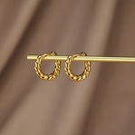 Layla Gold Twist Earrings, pair of twisted gold hoops hanging on a gold rod against brown fabric