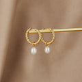 Layla Pearl Twist Earrings, pair of twisted gold hoops with pearl drops, resting on a gold bar