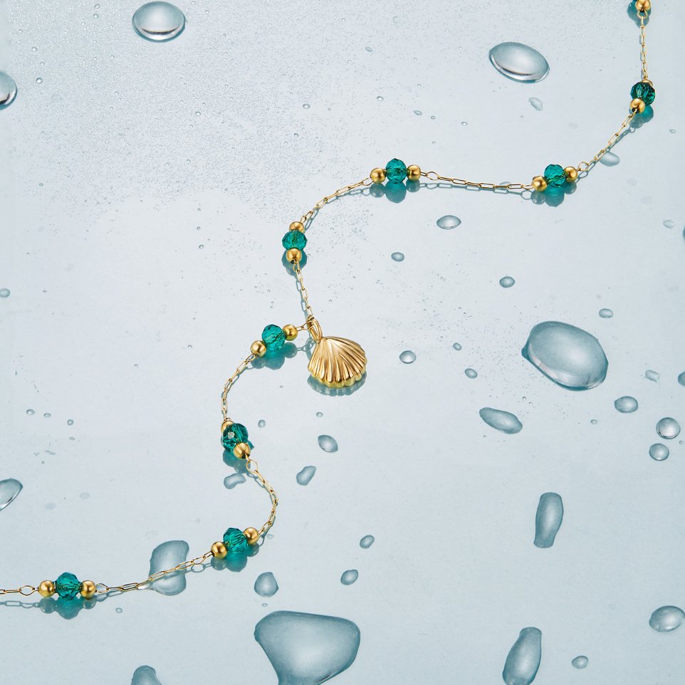 Nerissa Gold Shell & Green Bead Necklace, laid across wet blue glass with a gold shell charm
