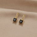Piper Black Crystal Drop Earrings, a pair of gold earrings with square black stones on beige
