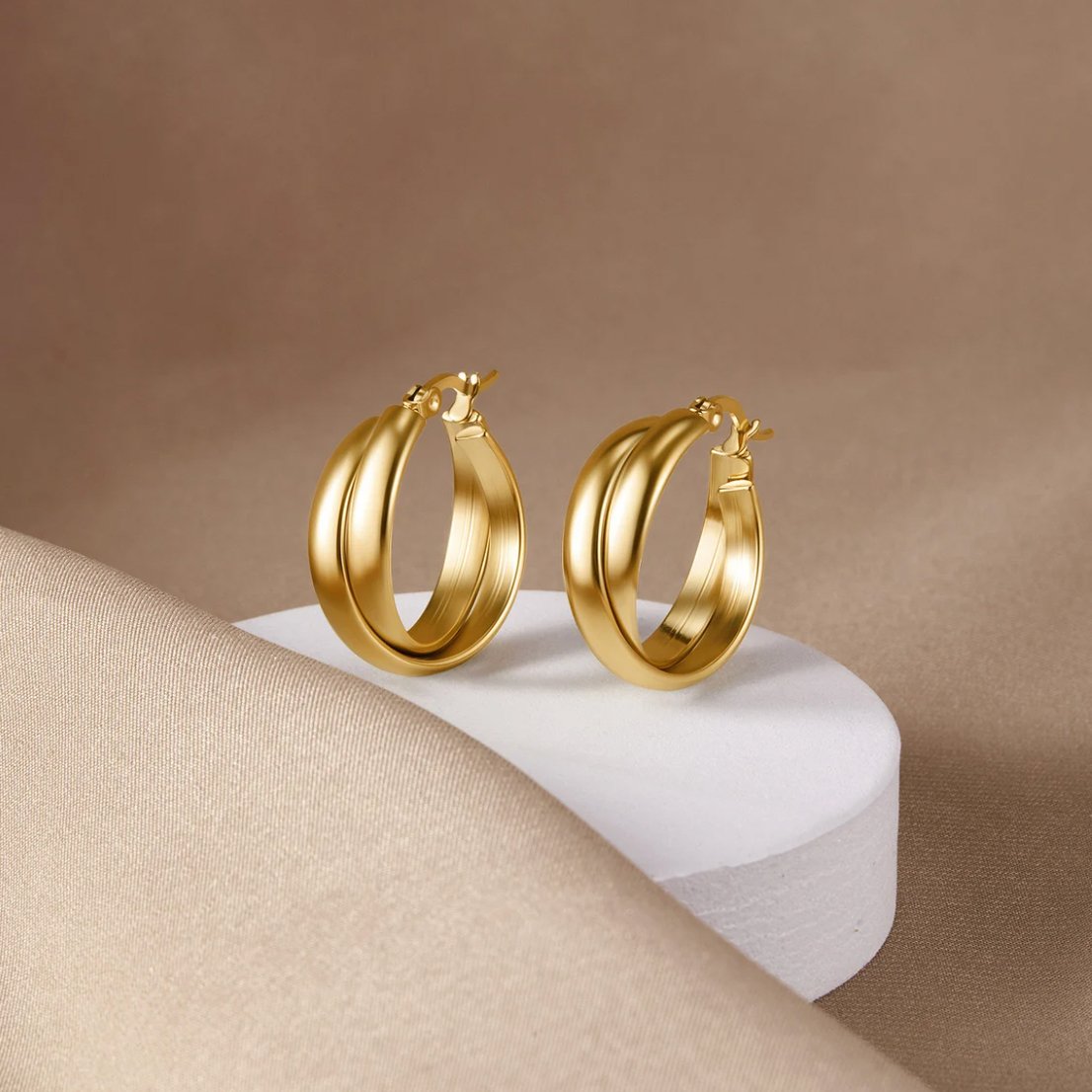 Talia Gold Hoop Earrings, a pair of gold hoops on a white pedestal over beige fabric