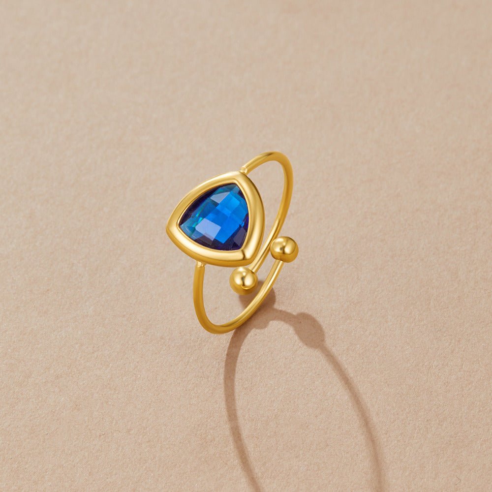 Via Sapphire Blue Ring, lying flat on sand-coloured surface, triangular blue stone